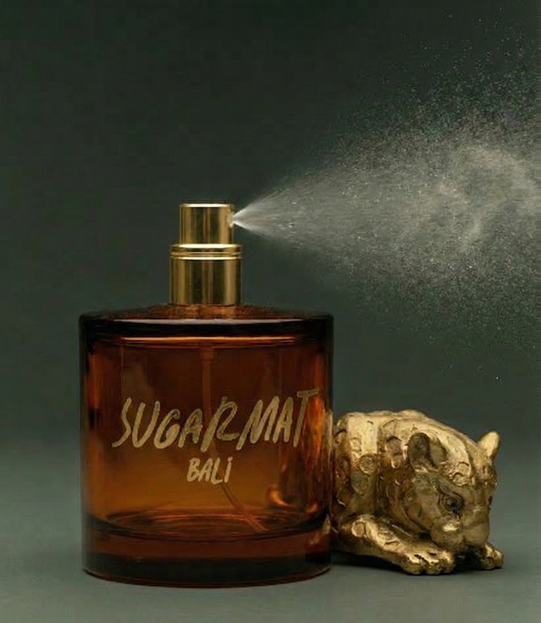 Brown perfume bottle labeled 'SUGAR MAT BALI' with gold spray nozzle and decorative figurine on a dark background
