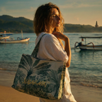 Woman holding a tote bag with a palm leaf design on a beach with boats and mountains in the background.