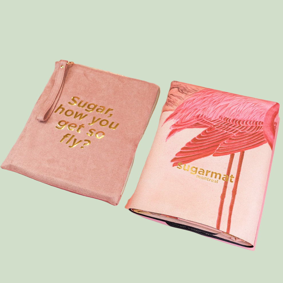 A suede yoga mat with a suede top layer and a natural rubber base, featuring a pink flamingo design and the text 'SUGAR, HOW YOU GET SO FLY?'. The mat appears to be rolled up at the top and placed on a pink surface.